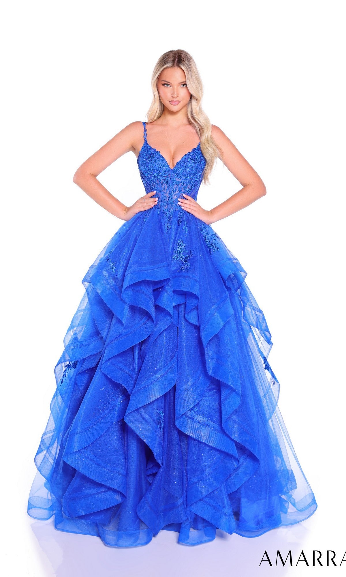 A woman wears the Amarra 89511 long prom dress, featuring a vibrant blue color, glitter tulle skirt, and an embellished fitted bodice, standing against a plain white background.